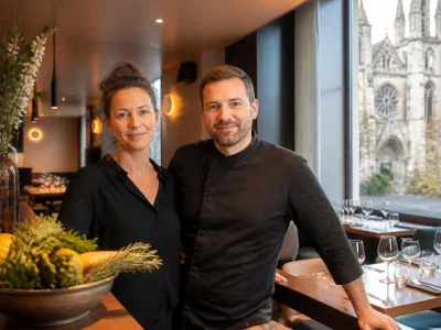 Un couple de restaurateurs souriants dans leur établissement situé au coins de la rue avec une église en arrière-plan
