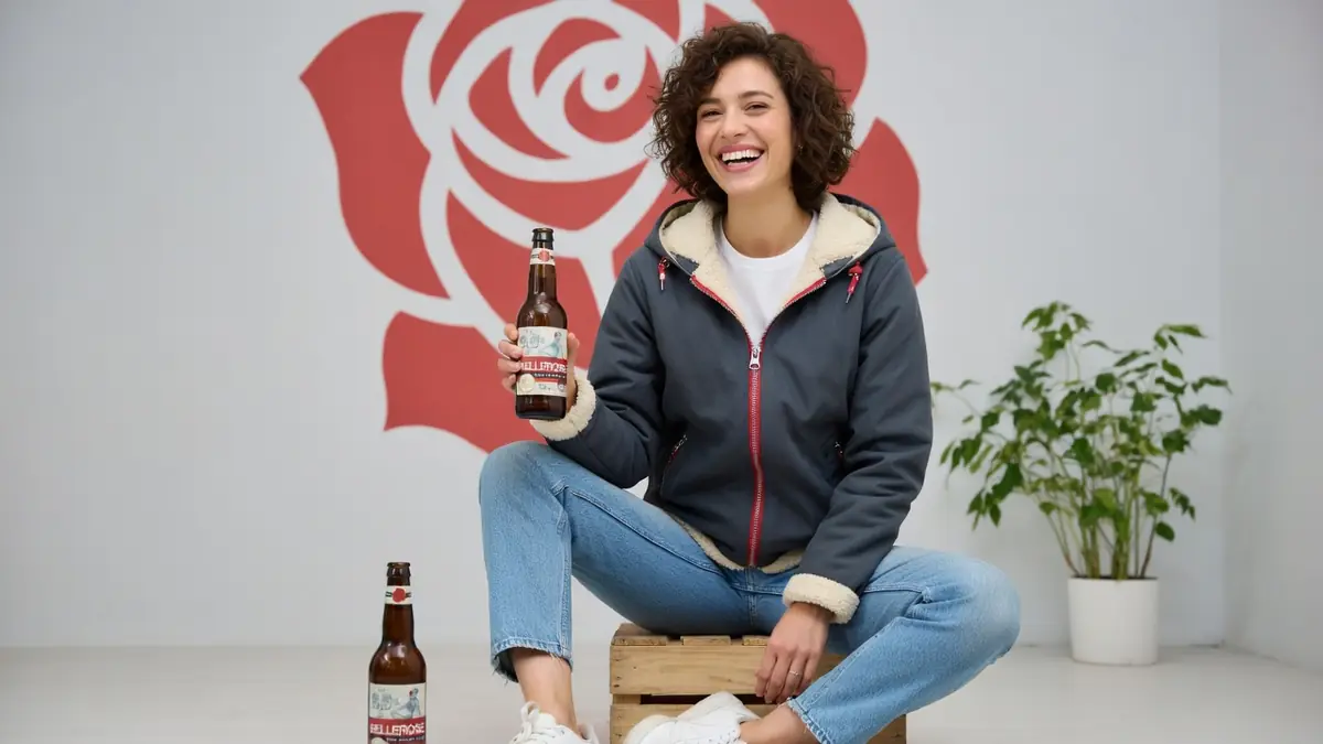 Une femme aux cheveux frisés sourit en tenant une bouteille tout en portant une veste Bellerose