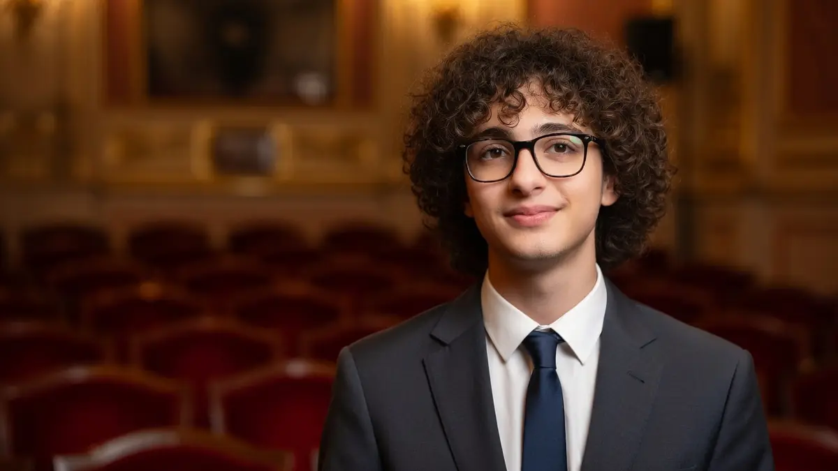 Portrait de Gabriel Naccache portant un costume et des lunettes dans une salle de théâtre aux sièges rouges