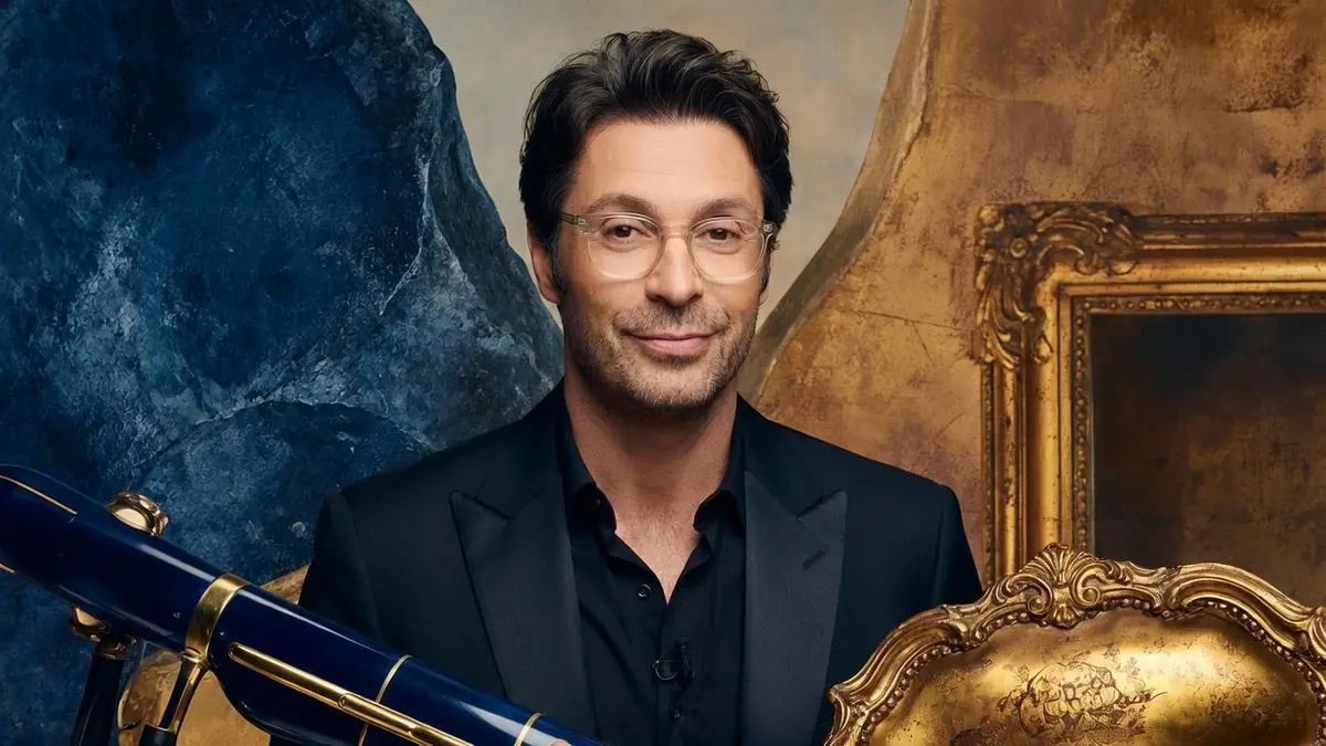 Johan Ledoux en costume noir sourit en tenant une guitare électrique bleue devant un décor baroque doré