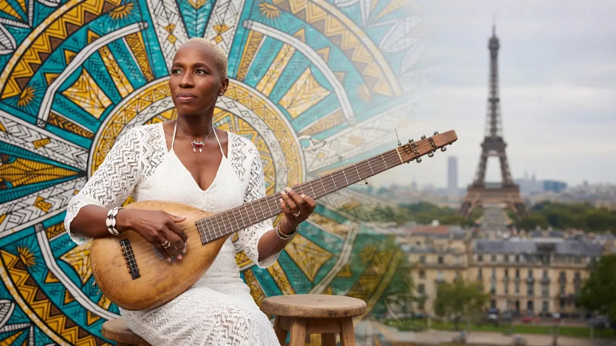 Kadi Diarra joue d'un instrument traditionnel devant un motif coloré et la tour Eiffel en arrière-plan