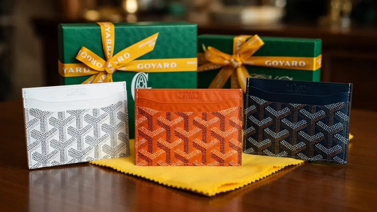 Plusieurs Goyard porte-carte colorés sont posés sur un tissu jaune devant des boîtes vertes