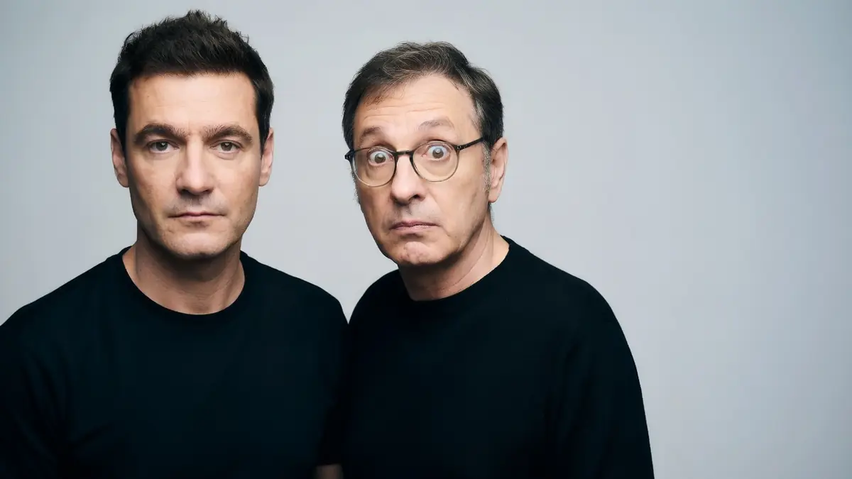 Manu Joucla pose avec un autre homme, l'un sérieux et l'autre faisant une grimace