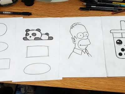Plusieurs feuilles de papier posées sur un bureau présentent des exemples de dessin facile comme un panda ou un personnage