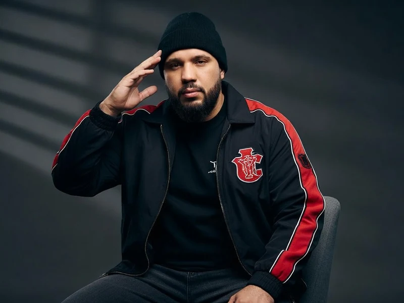 Kofs est assis avec une main près de la tête et une veste noire à bandes rouges