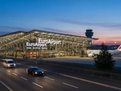 Des voitures circulent devant le terminal de l'aéroport de Bâle-Mulhouse avec un avion sur le tarmac au crépuscule
