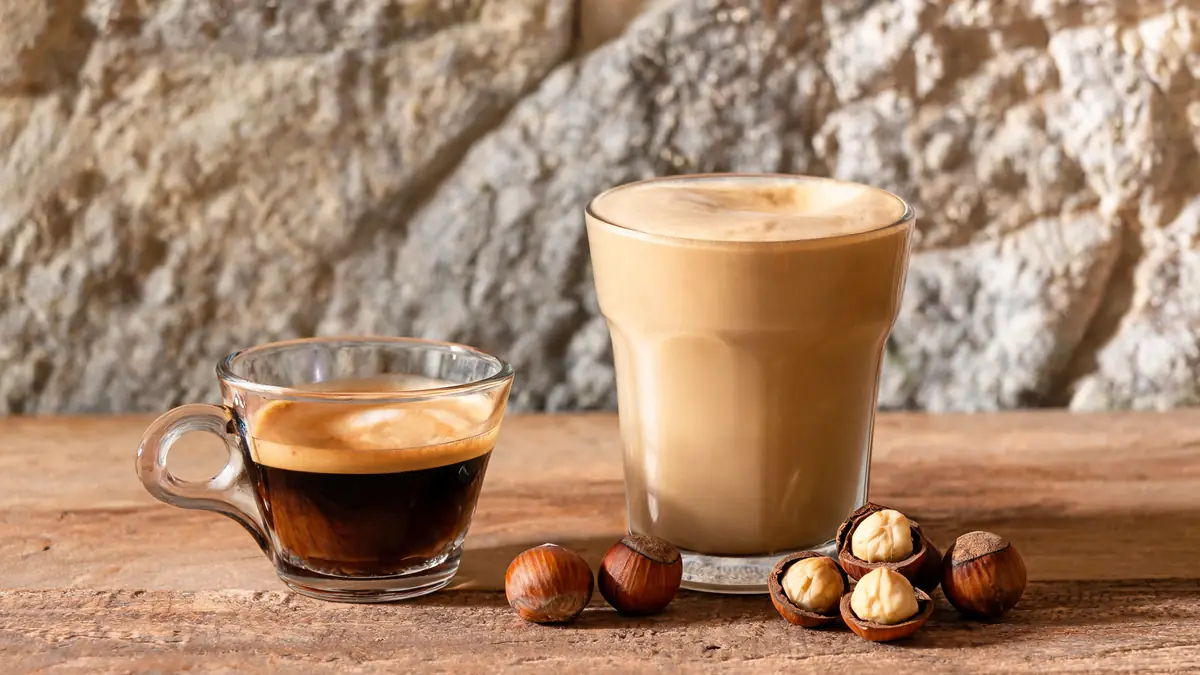 Deux verres de café dont un café à la noisette accompagnés de noisettes sur une table en bois