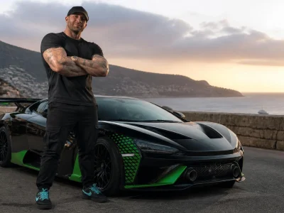 Gmk taille impressionnante à côté de sa supercar noire et verte sur une route côtière au coucher du soleil