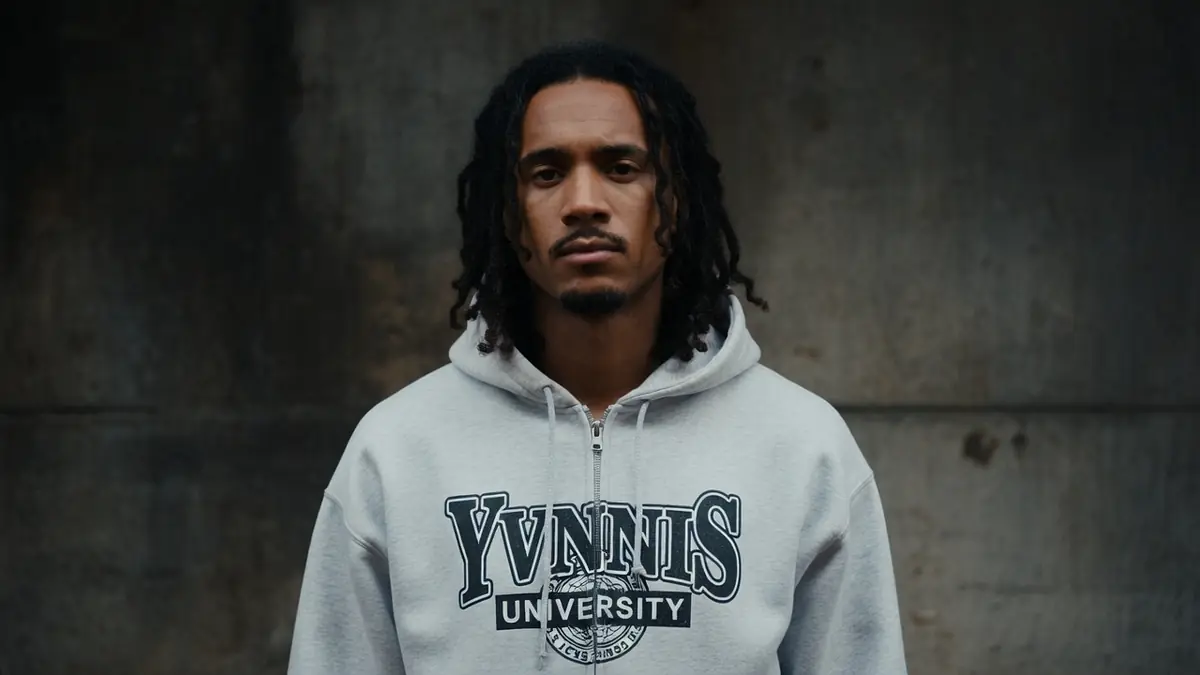 Yvnnis se tient debout en sweat gris devant un mur en béton