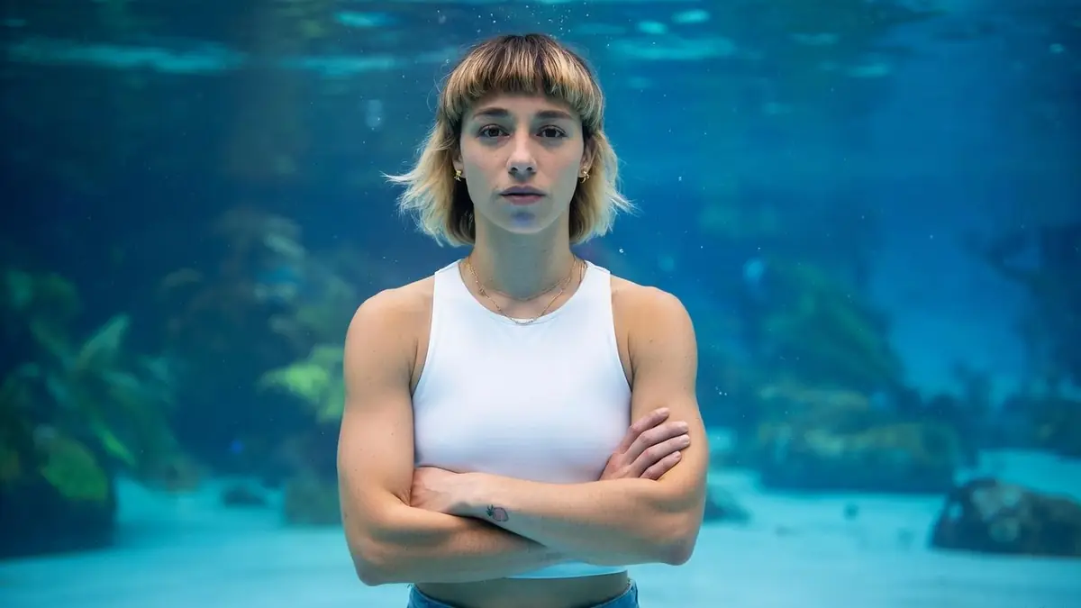Léa Catania pose en tenue blanche sous l'eau avec les bras croisés devant un décor aquatique