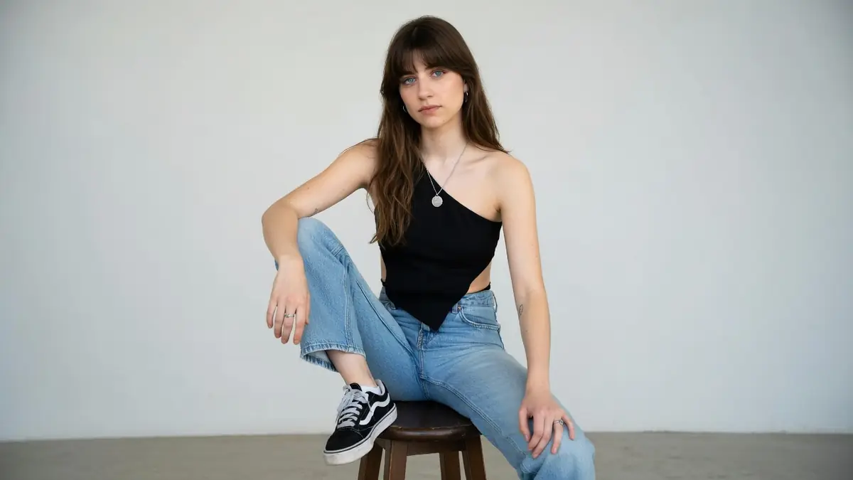 Lola Klodawski assise sur un tabouret portant un haut noir asymétrique et un jean bleu
