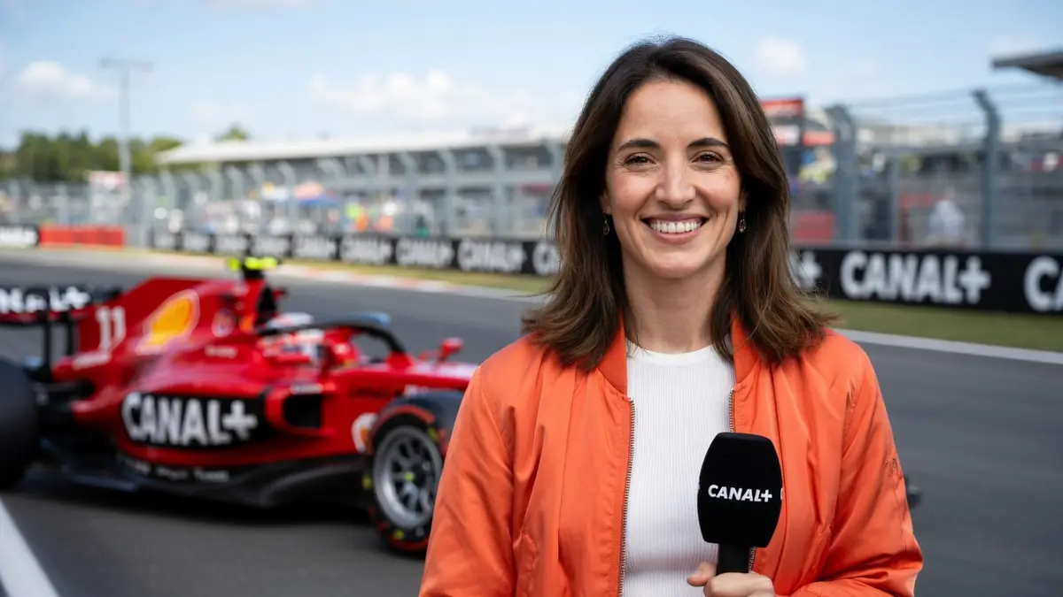 Margot Laffite sourit sur la piste d'un circuit avec une voiture de Formule 1 rouge en arrière-plan