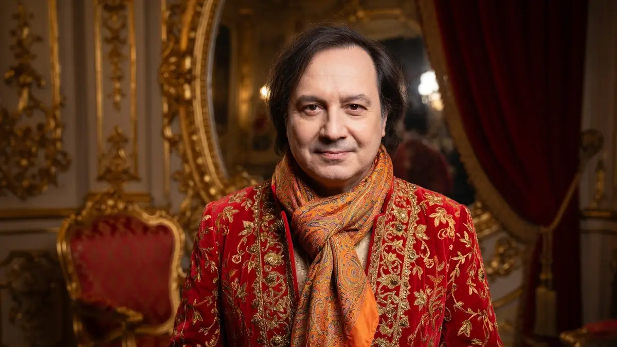 Michel Fau se tient dans un salon doré avec une veste rouge brodée et une écharpe colorée