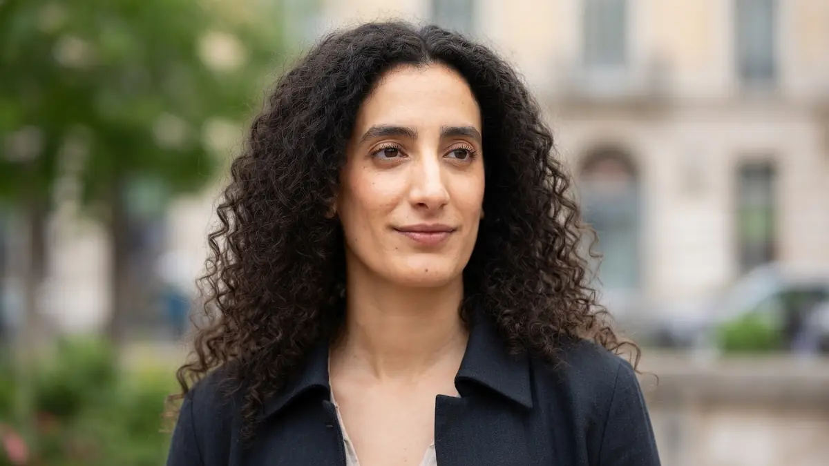 Portrait de Mouna Soualem avec ses longs cheveux bruns bouclés devant un arrière-plan urbain flou
