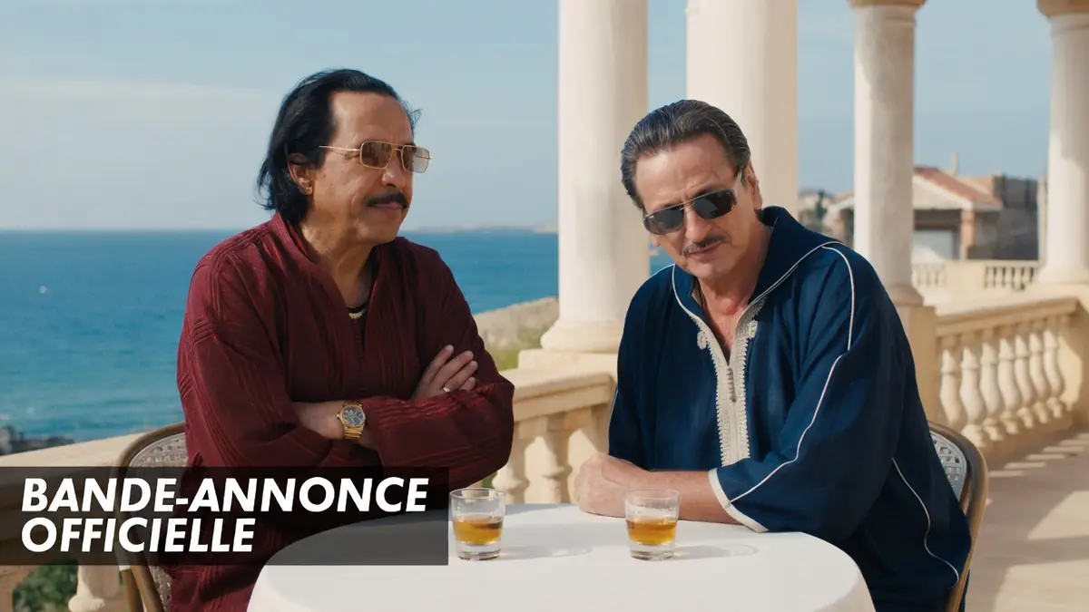 Deux hommes en djellabas et lunettes de soleil discutent à une table face à la mer dans le film omar La Fraise
