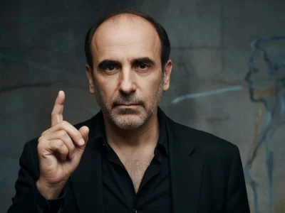 Philippe Harel lève l'index dans un portrait sombre en veste noire sur fond gris
