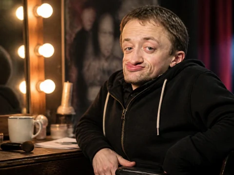 L'humoriste Guillaume Bats assis devant un miroir de loge avec un éclairage de scène en arrière-plan