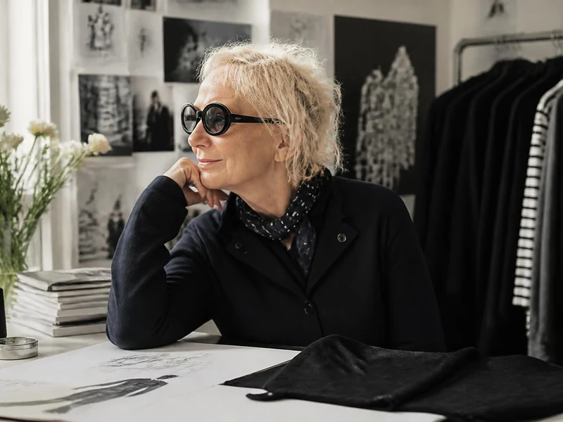 Portrait d'Agnès Troublé assise à son bureau, portant des lunettes de soleil et réfléchissant devant ses croquis de mode