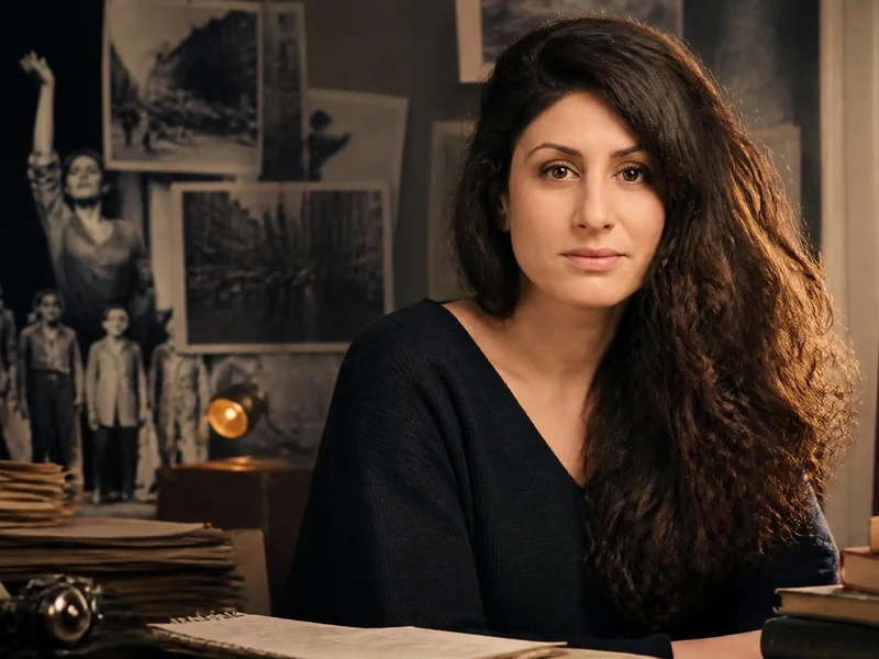 Portrait de l'autrice et comédienne Aïda Asgharzadeh posant devant un bureau rempli de documents et de livres