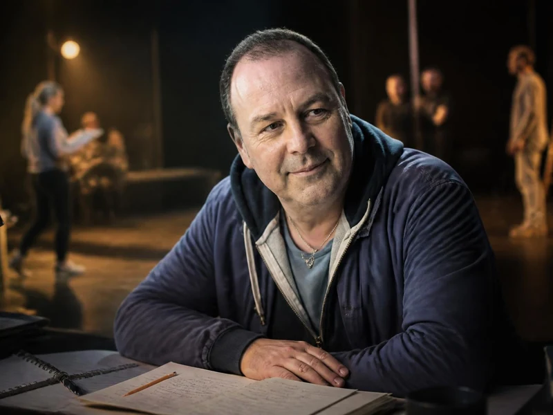 Portrait de Christophe Rauck assis à une table de travail avec des comédiens en répétition en arrière-plan