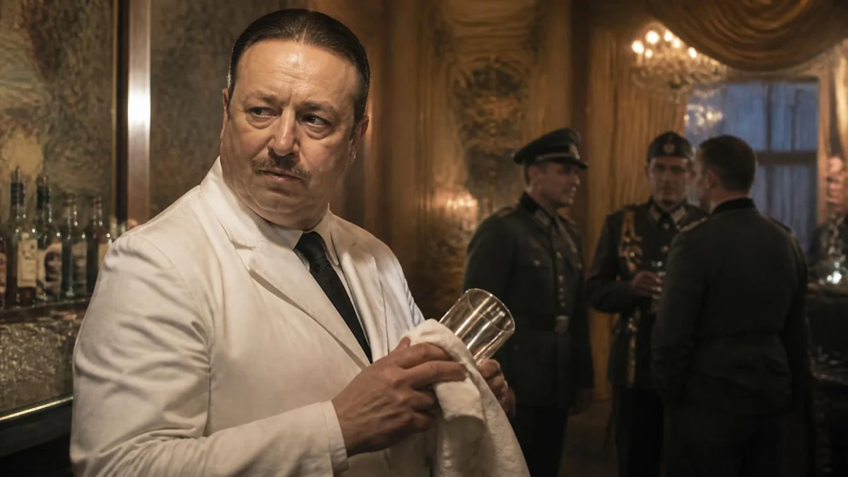 Un barman en veste blanche essuie un verre, incarnant Frank Meier au milieu d'officiers allemands dans un bar chic