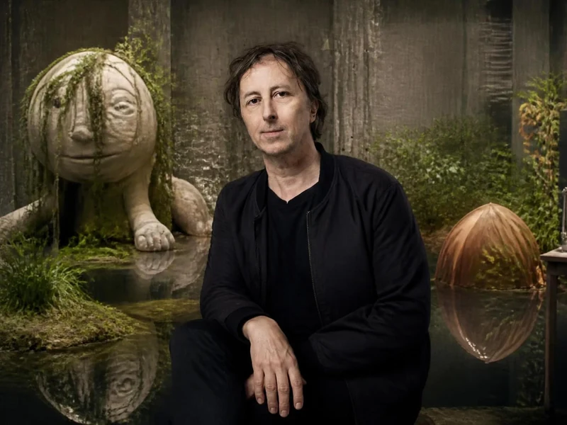 Portrait de Philippe Quesne posant devant un décor théâtral onirique peuplé de sculptures étranges et de végétation