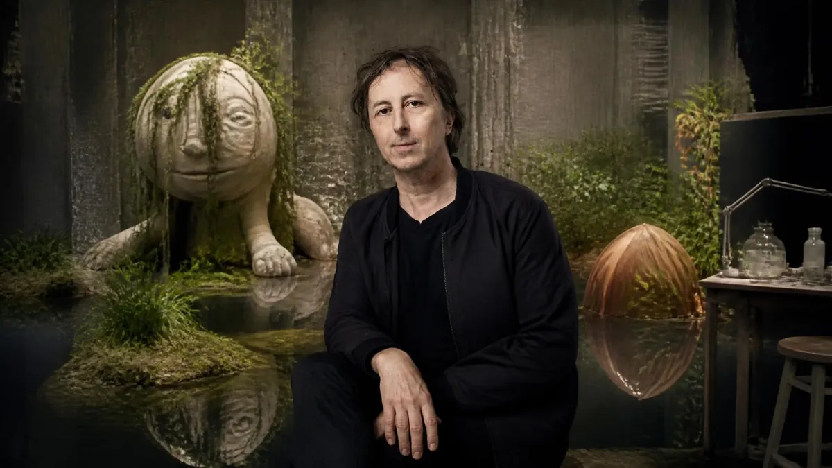 Portrait de Philippe Quesne posant devant un décor théâtral onirique peuplé de sculptures étranges et de végétation